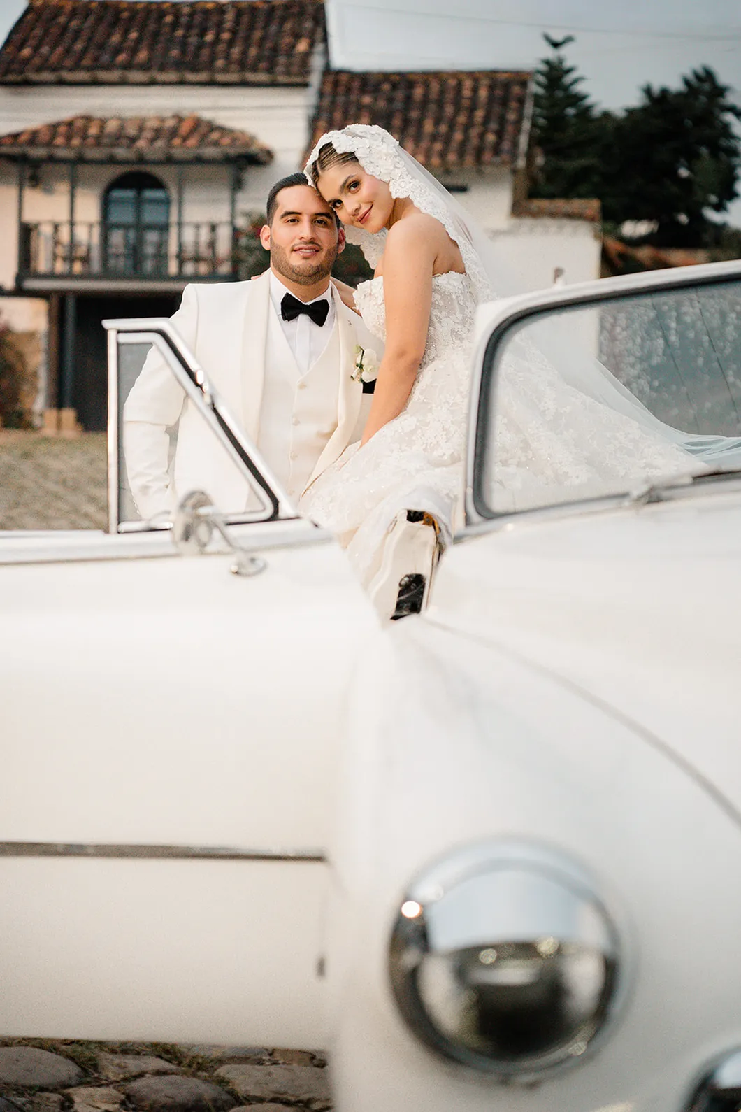 Los novios apoyados en un coche clasico blanco frente a una casa colonial, en Cartagena, por Omar Fraija
